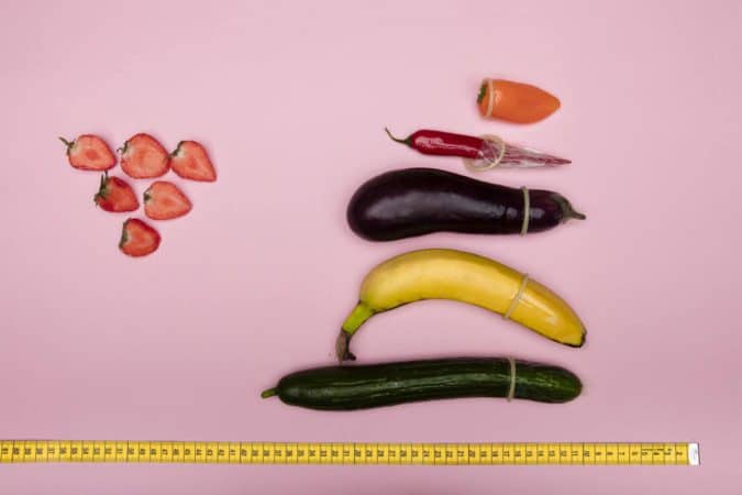 O objetivo da Omens é dar dicas sobre como saber o tamanho da camisinha . A imagem mostra um pimentão laranja, uma pimenta vermelha, uma berinjela, uma banana e um pepino de vários tamanhos embrulhados num preservativo em um fundo rosa.