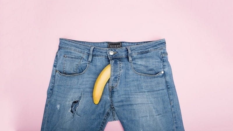 Uma banana caída, saindo pelo zíper da calça jeans, representando uma ereção fraca