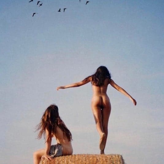duas mulheres nuas ao ar livre, uma sentada, outra de pé com os braços abertos
